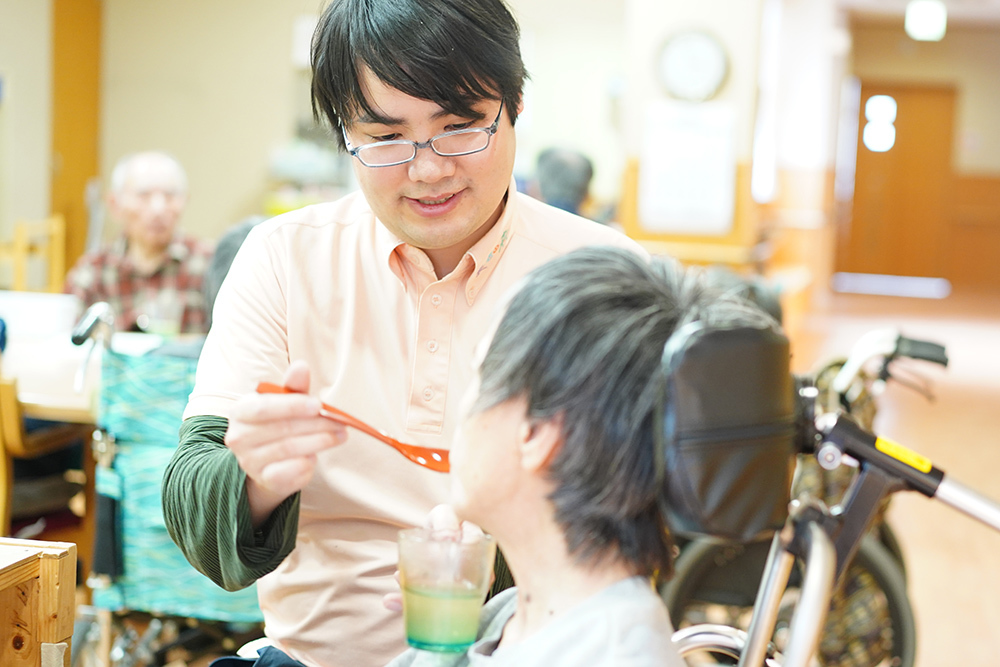 スタッフが入居者に食事介助を行っている風景。明るい共用スペースでの支援場面。